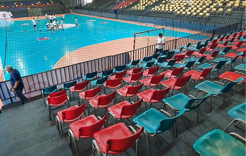 En la imagen vemos la instalación deportiva del Centro de Entrenamiento Olímpico de Ñuñoa desde la gradería central. Allí hay sillas rojas y azules, y se aprecia el perímetro del gimnasio que, en su centro, tiene a un grupo de 10 personas entrenando.
                                