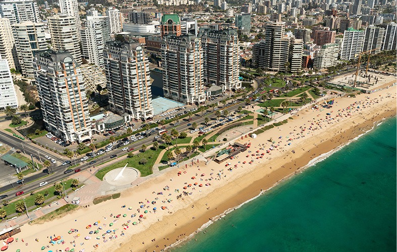 On the image, an aerial shot of the coast of Viña del Mar 
                            is appreciated. On the inferior left area is the sea with its 
                            waves. In the middle is the beach and green areas. At 
                            the back there are high-rise buildings.